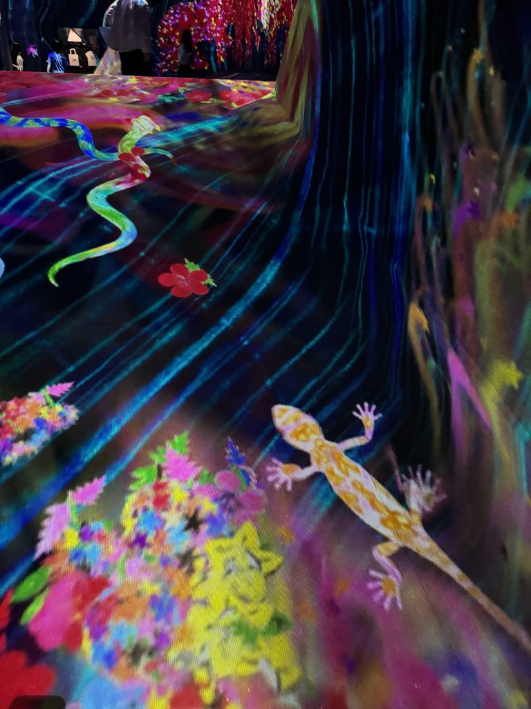 A digital gecko crawling across a floor of projected flowers at teamLab