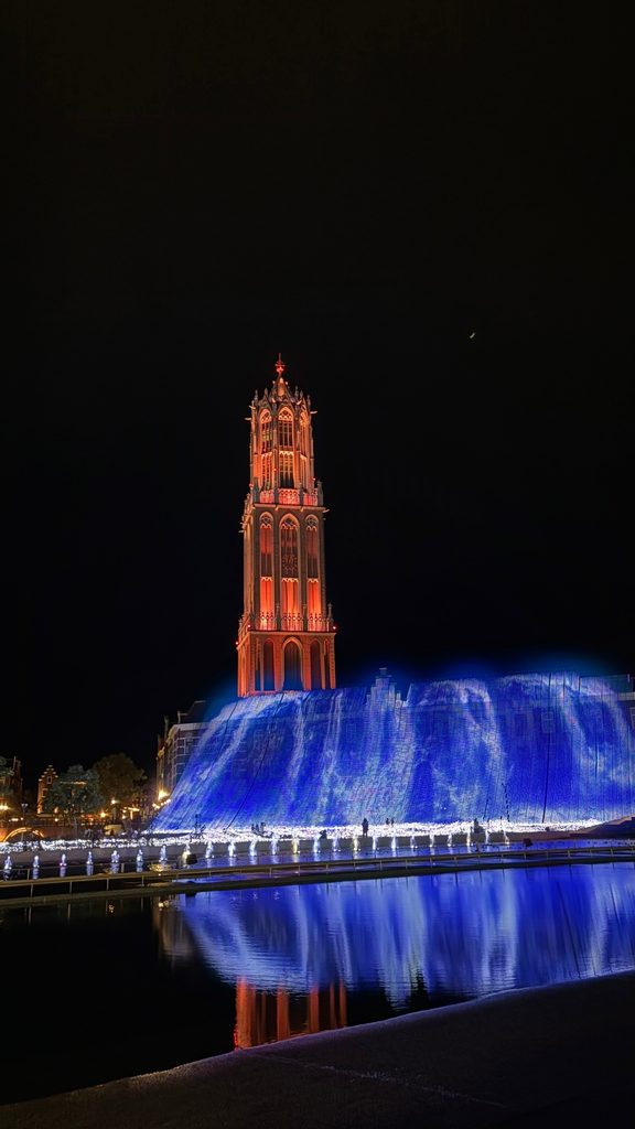 The Domtoren tower glowing red above a blue-lit fountain, mirrored in the water at Huis Ten Bosch
