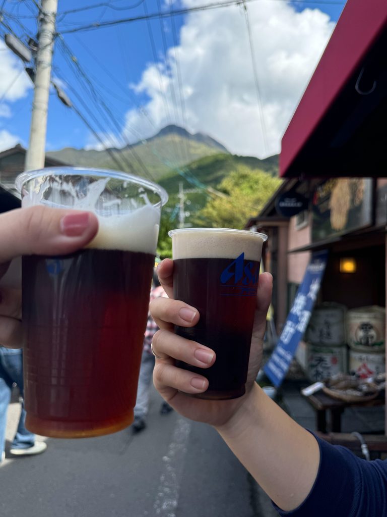 Toasting with two cups of local dark beer on Yunotsubo Street, with Mt. Yufu in the background